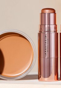 Twee beige cosmetische producten: een ronde, gladde crème in een doorzichtige pot en een metallic stick highlighter met een sprankelende top, beide in roze verpakkingen.