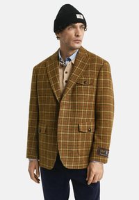 Mann in einem braunen karierten Blazer über einem beigefarbenen Hemd mit Knöpfen und einem blauen Poloshirt, mit marineblauen Hosen und einer schwarzen Strickmütze, schaut nach rechts.