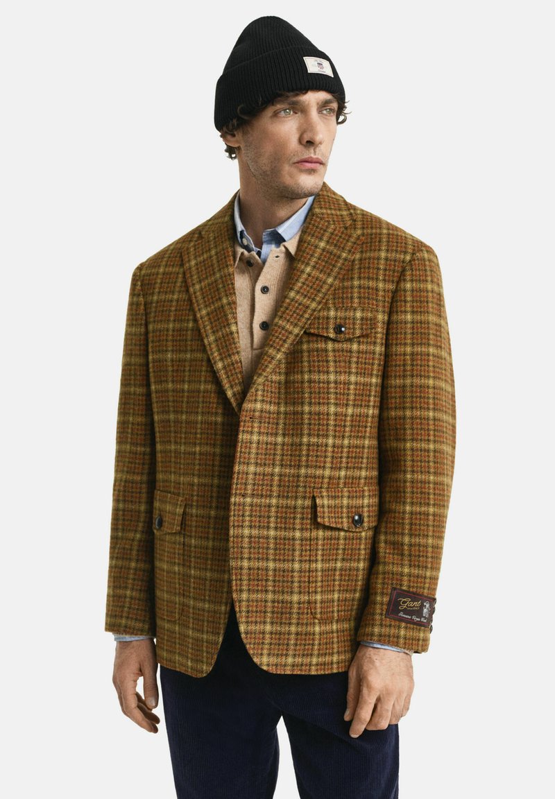 Mann in einem braunen karierten Blazer über einem beigefarbenen Hemd mit Knöpfen und einem blauen Poloshirt, mit marineblauen Hosen und einer schwarzen Strickmütze, schaut nach rechts.