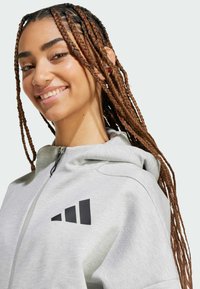 Γκρι φούτερ ζιπ με υφή, επενδεδυμένη κουκούλα και έντονο μαύρο λογότυπο στο στήθος. Λείος τελικός φινίρισμα με πλεξούδα μαλλιών.