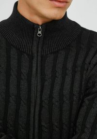 Black knit zip-up sweater with a ribbed collar, featuring textured cable patterns and a smooth zipper, combining different shades of black.