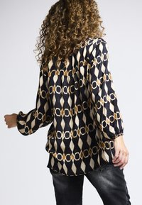 Langärmelige schwarze Bluse mit geometrischem Muster in goldenen, weißen und braunen Tönen. Verfügt über einen gerafften Rücken und elastische Bündchen.