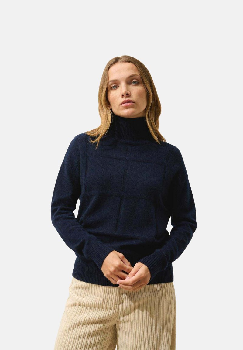 Maglione a collo alto blu navy con un motivo a griglia testurizzato, polsini e orlo a coste. Indossato con pantaloni a coste di colore chiaro.