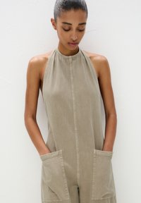 Beige Jumpsuit mit hohem Kragen, vorderer Knopfleiste und großen Vordertaschen. Der Stoff hat eine glatte, weiche Textur.