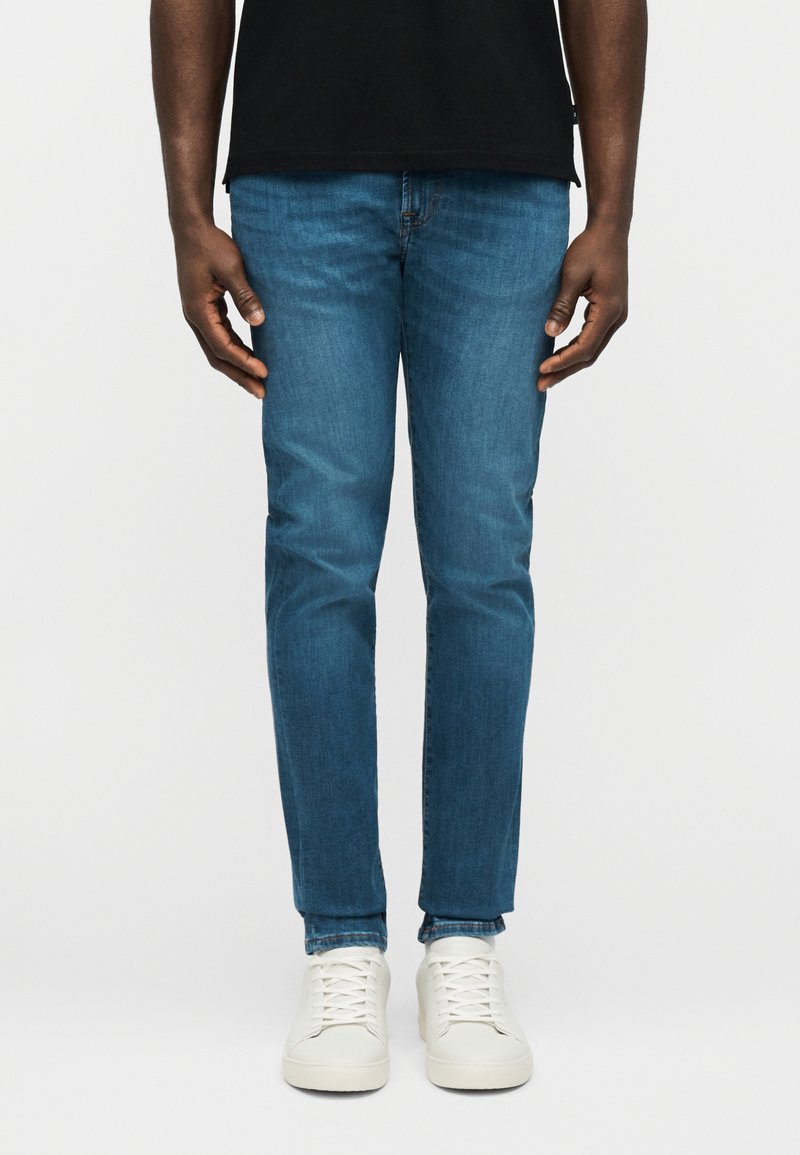 Άτομο που φορά στενά μπλε τζιν, λευ κά αθλητικά παπούτσια και μαύρο πουκάμισο, στέκεται μπροστά σε απλό λευκό φόντο.