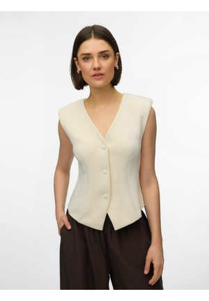 Vrouw met kort bruin haar, gekleed in een mouwloos crème vest met knopen en losse donkerbruine broek, tegen een effen witte achtergrond.