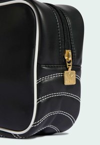 Czarna skórzana torba z białymi lamówkami, zakrzywionym białym przeszyciem oraz złotym suwakiem z tłoczonym logo. Kompaktowy i prostokątny kształt.