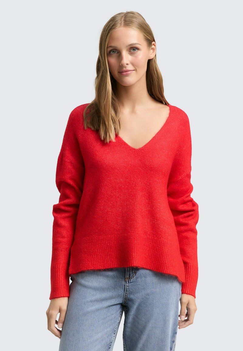 Rode V-hals trui met lange mouwen en geribbelde zoom, gemaakt van een zachte, gestructureerde stof. Gepaard met lichtblauwe jeans.