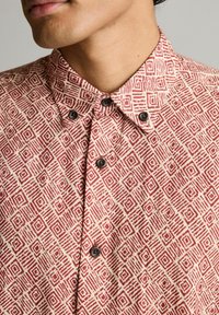Chemise à motifs rouge et crème avec un design géométrique, dotée d'un col pointu et de boutons noirs. Tissu texturé avec un léger éclat.