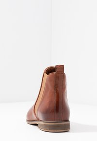 Brune lær ankle boot med elastiske sidepaneler, glat tekstur og en lav stablet hæl. Designet har stikninger som accent.