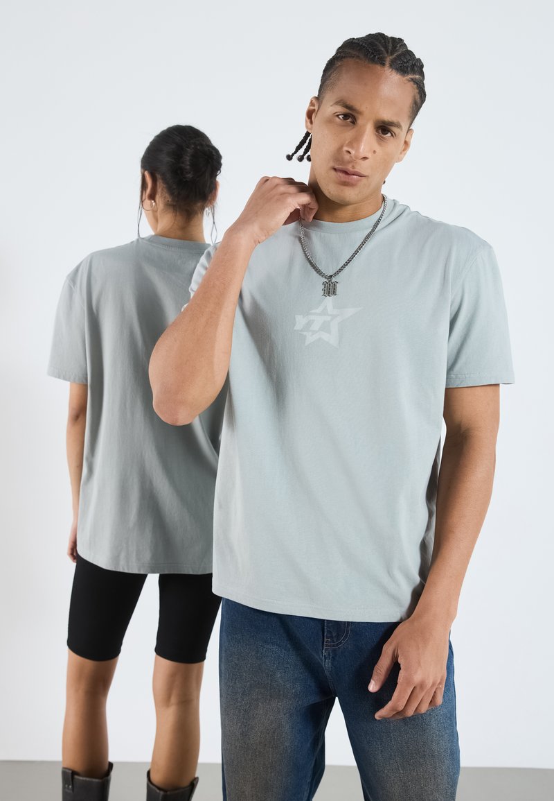 Camiseta de gola redonda cinza claro com um discreto logótipo de estrela na parte da frente, combinada com jeans azuis. O tecido parece macio e casual.