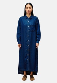 Lange denim shirtjurk met kraag, doorgeknoopte voorkant, borstzak en lange mouwen. Diepblauwe kleur, rechtstaande pasvorm en zijsplits aan de zoom.