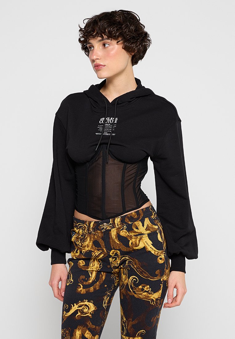 Sudadera negra recortada con capucha y cremallera frontal, superpuesta a un corsé transparente. Combinada con pantalones negros y dorados con estampado.