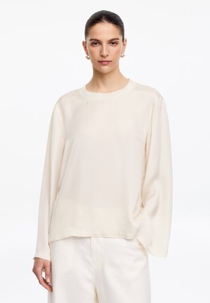 Femme aux cheveux foncés tirés en arrière, portant des boucles d'oreilles en perles, un chemisier ample crème à manches longues et un pantalon blanc sur fond blanc.