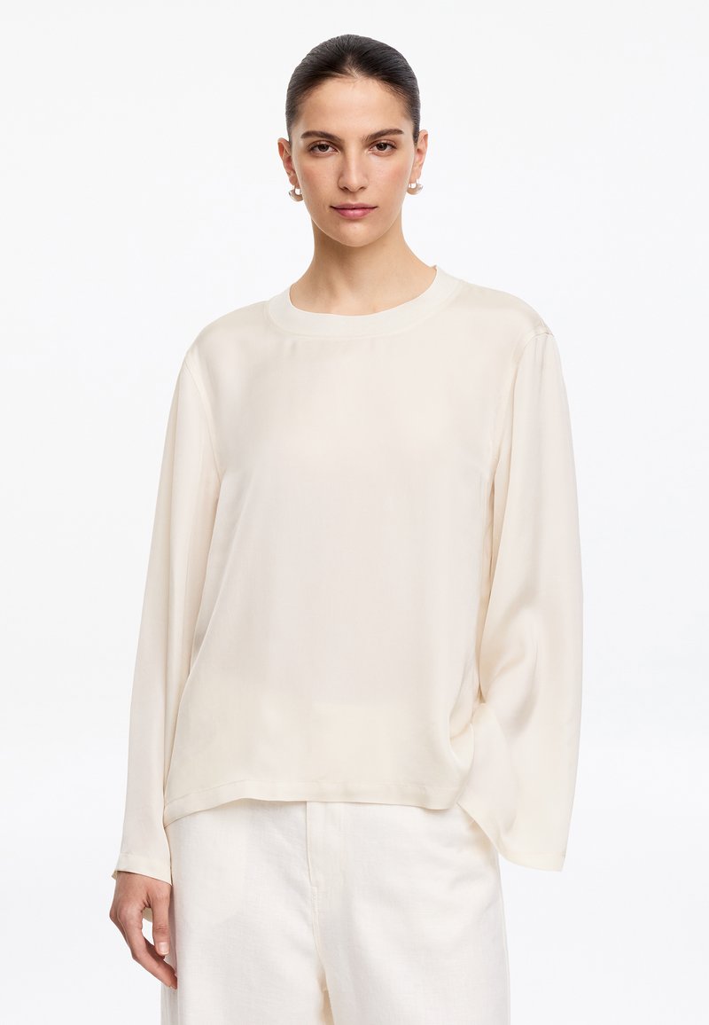 Femme aux cheveux foncés tirés en arrière, portant des boucles d'oreilles en perles, un chemisier ample crème à manches longues et un pantalon blanc sur fond blanc.
