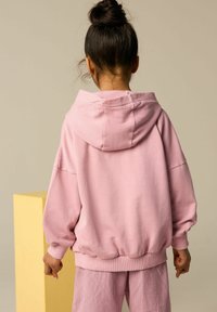 Różowa bluza z kapturem o luźnym kroju, opadających ramionach i ściągaczach. Noszona z dopasowanymi, fakturowanym różowymi spodniami, pokazana z tyłu.