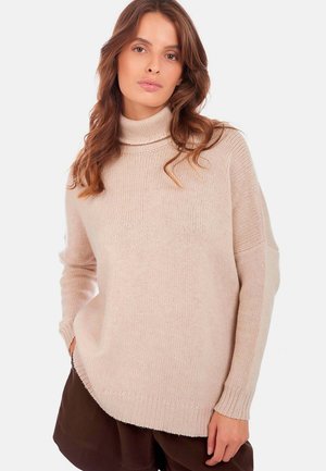 Pull à col roulé beige clair avec une coupe décontractée, tricot texturé, manches longues et ourlet côtelé. Porté par-dessus un short marron foncé.