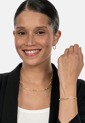 Gold jewelry set featuring a linked necklace and bracelet with textured accents, worn over a white top and black blazer.