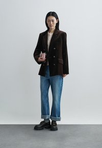 Femeie în picioare purtând un sacou maro închis cu două rânduri de nasturi, pulover deschis la culoare, blugi albastri suflecați, încălțăminte neagră masivă, ținând o poșetă roz mică.