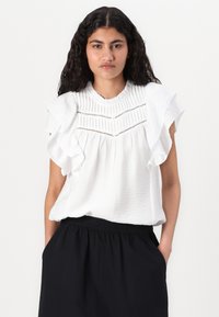Haut texturé blanc avec des manches volantes, présentant un col plissé et des coupures horizontales, associé à une jupe noire à taille élastique.