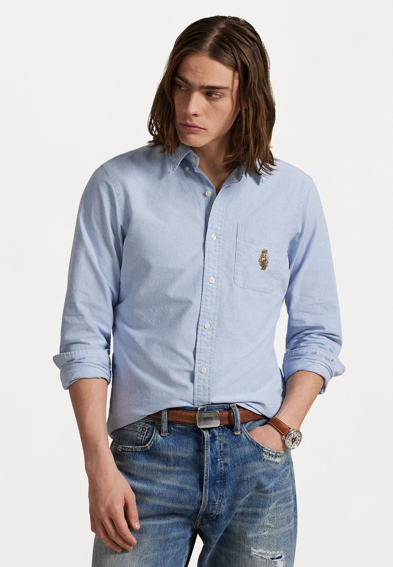 Lichtblauw overhemd met lange mouwen, borstzak en geborduurd ontwerp. Draagt het met versleten jeans en een bruine riem.