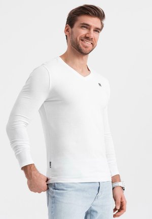 Langärmliges, weißes V-Ausschnitt-Shirt mit kleinem Logo auf der Brust, aus einem glatten, leichten Stoff, kombiniert mit hellblauen Jeans.