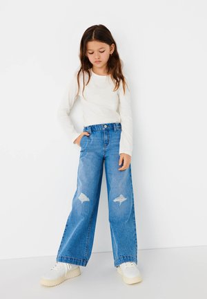 Jeans en denim bleu à jambes larges avec des patches déchirés, associés à une chemise blanche à manches longues et des baskets blanches. Look simple et décontracté.