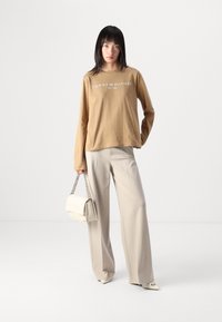 Langärmliges, sandfarbenes T-Shirt mit weißem Logotext, kombiniert mit hellbeigen, weit geschnittenen Hosen und einer cremefarbenen Handtasche mit Kettenriemen.