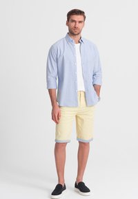 Camicia azzurra chiaro con maniche arrotolate, t-shirt bianca sotto, abbinata a pantaloni corti gialli e scarpe slip-on nere.