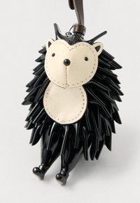 Breloc în formă de arici, cu fața bej și corp negru cu țepi, atârnând de o curea maro din piele pe un fundal alb.