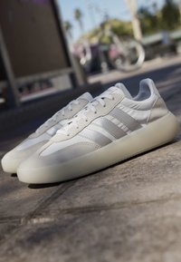Paire de baskets Adidas blanches et gris clair avec semelles épaisses translucides, placée à l'extérieur sur un trottoir près d'un vélo flou.