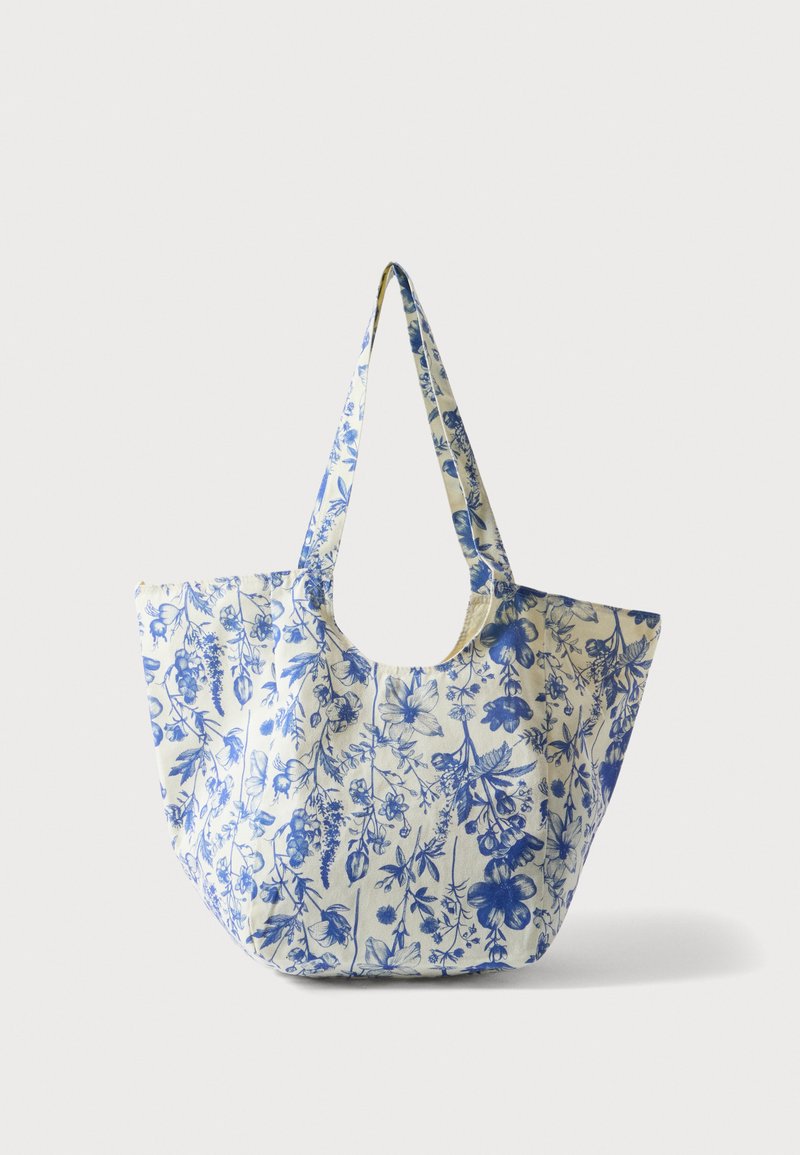 Grand sac cabas crème avec imprimé floral bleu détaillé et larges bretelles, posé sur un fond blanc.