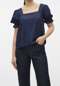 Marinblus med kort ärm i strukturerad tyg med fyrkantig halsringning och puffärmar, kombinerad med mörka denimbyxor.