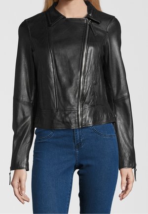 Frau trägt eine schwarze Lederjacke mit diagonalem Frontreißverschluss und blaue Jeans, steht vor einfarbigem Hintergrund.