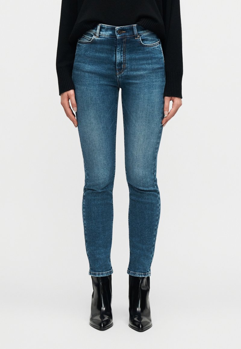 Γυναίκα που φοράει εφαρμοστά μπλε  τζιν, μαύρα μυτερά μποτάκια αστραγάλου και μαύρο πουλόβερ με μακριά μανίκια, στέκεται μπροστά σε ένα απλό λευκό φόντο.