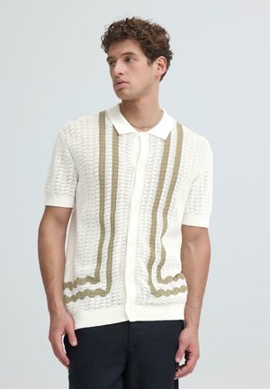 Mladi moški s kodrastimi lasmi, oblečen v belo pleteno majico s kratkimi rokavi z bež navpičnimi črtami, gleda v stran na enobarvnem ozadju.