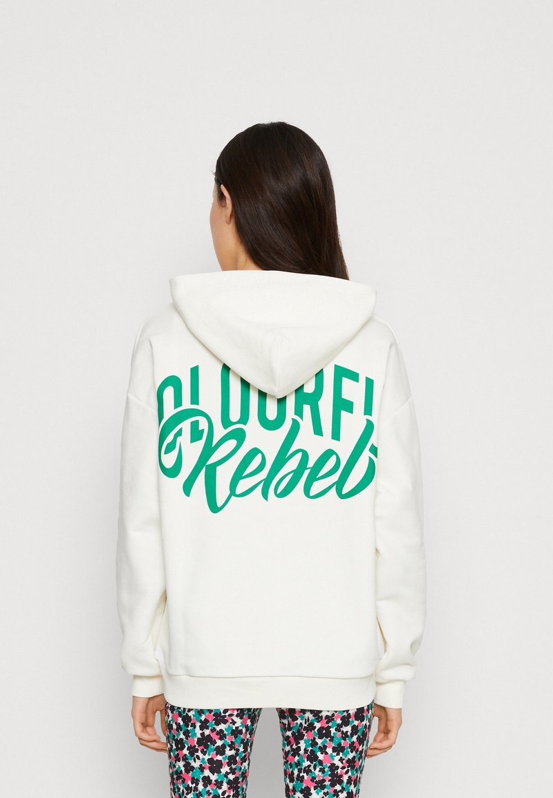 Felpa bianca con il testo verde "Colorful Rebel" sulla schiena, caratterizzata da un ampio cappuccio e una vestibilità comoda. Abbinata a leggings con un motivo floreale.