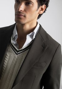 Joven con un blazer oscuro sobre un suéter de punto en color crema y una camisa de vestir blanca a rayas, mirando ligeramente de lado.