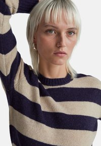 Kobieta o krótkich blond włosach, ubrana w granatowo-beżowy sweter w paski, unosząca jedną rękę za głowę, o neutralnym wyrazie twarzy, na białym tle.