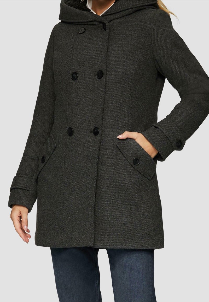 Femme portant un manteau gris foncé à double boutonnage avec de gros boutons, des détails aux poignets, et une main dans la poche latérale, associé à un jean bleu.