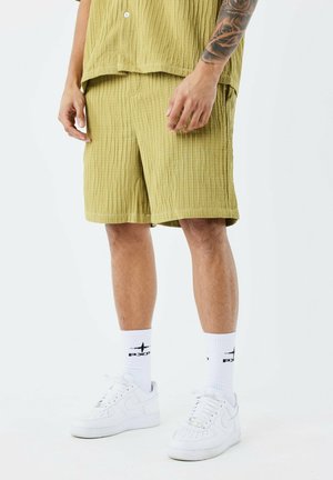 Shorts vert clair, texturés, avec une coupe décontractée, associés à des baskets blanches et des chaussettes mi-mollet. L'ensemble présente de légères rainures verticales.