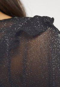 Tessuto nero trasparente con fine dettaglio di glitter argento, caratterizzato da un bordo arricciato sulla spalla, che mostra un motivo testurizzato sulla superficie.