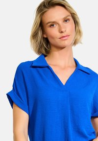 Modrá blouse s krátkým rukávem a límečkem ve tvaru V, vyrobená z texturované látky a s ohrnutými manžetami.