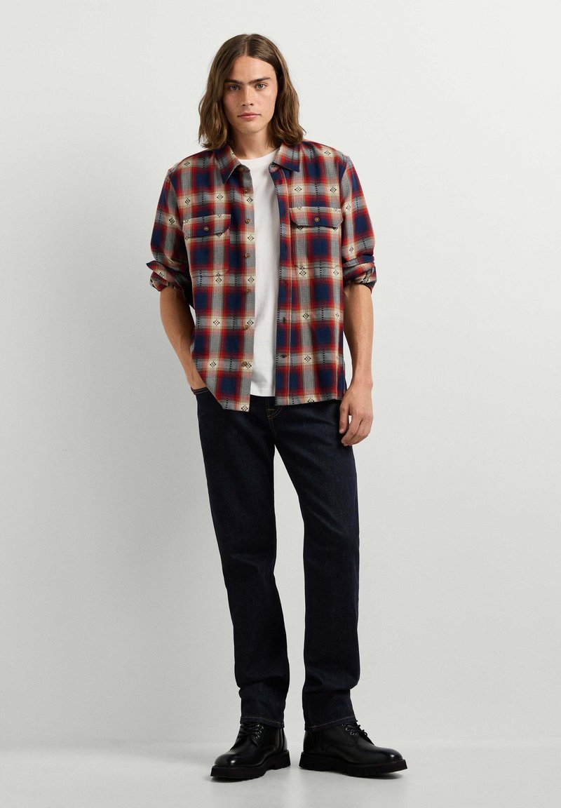 Homme aux cheveux bruns mi-longs portant une chemise à carreaux rouge et bleue sur un t-shirt blanc, un jean foncé et des chaussures noires, debout contre un mur blanc.