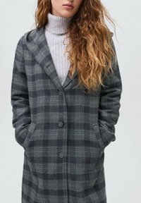 Kvinna klädd i en grå och svart rutig kappa över en ljusgrå polotröja, med händerna i fickorna på kappan och långt lockigt hår.