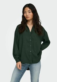 Dark green button-up shirt with a relaxed fit, long sleeves, and a shallow collar, made from a textured fabric. Worn with light blue jeans.