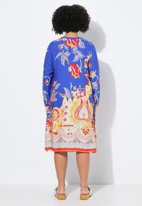 Farbenfrohes blaues Kleid mit floralen Mustern, langen Ärmeln, knielang, mit orangefarbenen und rosafarbene Akzenten am Saum. Flache Schuhe mit Holz-Effect Absatz.
