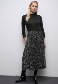 Vrouw in een zwarte aansluitende top met driekwartmouwen en een zwarte geplooide midi-jurk met een grijs abstract patroon, gecombineerd met zwarte enkellaarzen.
