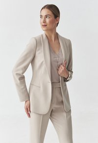 Vaatteellinen beige bleiseri, jossa on viistetty kaulus, yhdistetty samanväriseen beigeen puseroon ja housuihin. Tasainen pinta ja siistit linjat. Hopeiset korusteet.