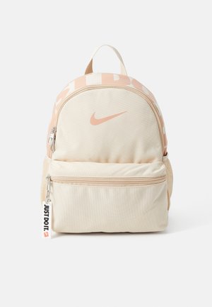 Mochila beige con acentos en rosa claro, que presenta un logo prominente de Nike, un bolsillo frontal con cremallera y una etiqueta tejida con el lema "JUST DO IT". Tejido texturizado.
